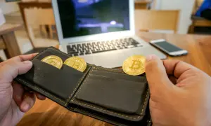 Ręka trzymająca portfel z trzema złotymi monetami bitcoin. Na stole leży laptop i smartfon. To symbol inwestycji na zonda giełda.