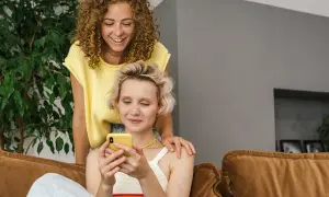 Dwie kobiety śmieją się, przeglądając telefon. Jedna z nich kładzie dłoń na ramieniu drugiej, sugerując wspólne konto dla par.