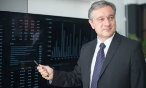 Mężczyzna w garniturze wskazuje na ekran z wykresami i danymi finansowymi.
