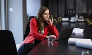 Kobieta w czerwonej marynarce pracuje przy laptopie przy długim stole konferencyjnym.