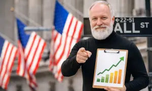 Starszy mężczyzna z brodą, wskazujący palcem, trzyma tablicę z wykresem wzrostu. Pokazuje, jak inwestować, na tle flag USA i znaku Wall Street.