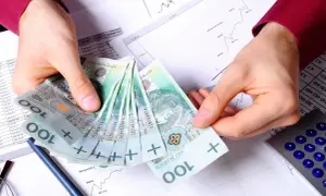 Dłonie trzymające stos polskich banknotów 100 zł na tle dokumentów finansowych i kalkulatora.