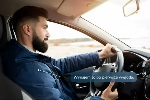 Mężczyzna z brodą prowadzi samochód, trzymając kierownicę.