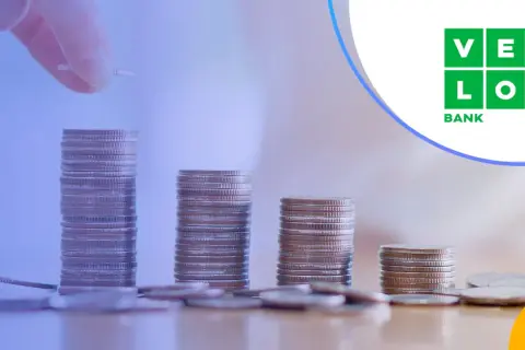 Ręka dodaje monetę do rosnącej stosu, symbolizując wzrost kapitału. VeloBank oprocentowanie lokat rośnie wraz z Twoimi oszczędnościami.