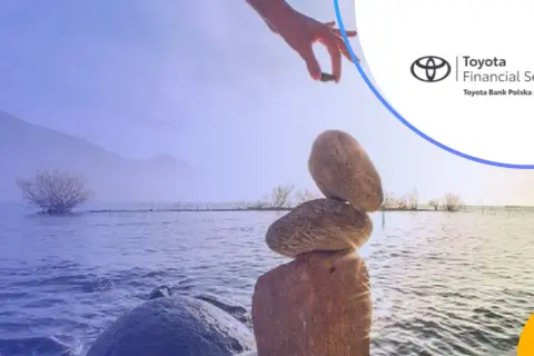 Równowaga kamieni na brzegu jeziora, ręka kładzie ostatni kamień. Logo Toyota Bank.