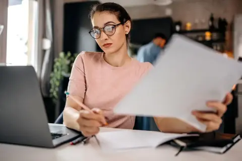 Kobieta w okularach analizuje dokumenty przy laptopie.
