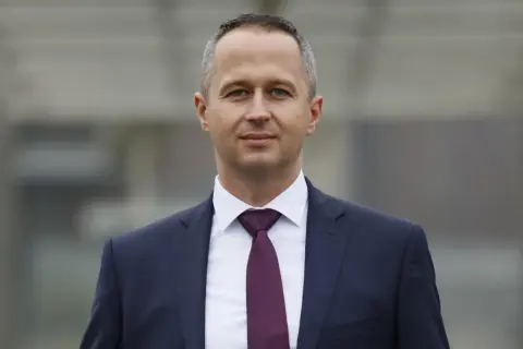 Mężczyzna w granatowym garniturze i bordowym krawacie, patrzący w obiektyw.