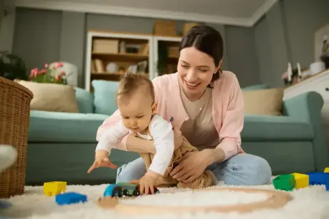 Uśmiechnięta mama bawi się z dzieckiem na dywanie, wśród kolorowych zabawek.