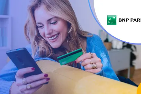 Uśmiechnięta kobieta z kartą płatniczą BNP Paribas w ręku korzysta z telefonu. Sprawdza BNP Paribas opłaty i prowizje.