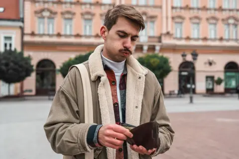 Młody mężczyzna z miną zmartwienia przegląda pusty portfel. W tle kamienice, symbolizujące inflację stylu życia.