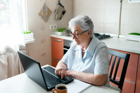 Starsza kobieta w okularach siedzi przy stole w kuchni i korzysta z laptopa.