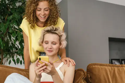 Dwie kobiety śmieją się, przeglądając telefon. Jedna z nich kładzie dłoń na ramieniu drugiej, sugerując wspólne konto dla par.