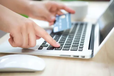 Dłoń z kartą zbliża się do klawiatury laptopa, by dokonać płatności. To może być rachunek rejestrowy PKO.