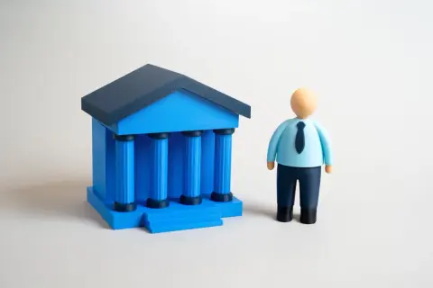 Figurka mężczyzny w niebieskiej koszuli i krawacie stoi obok niebieskiego budynku z kolumnami. Wibid on.