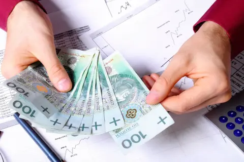 Dłonie trzymające stos polskich banknotów 100 zł na tle dokumentów finansowych i kalkulatora.