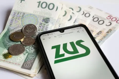 Złote banknoty i monety na nich leżący smartfon z logo ZUS.