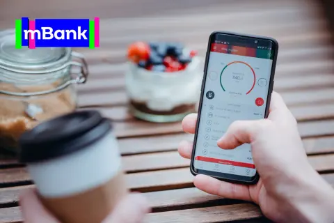 Dłoń trzyma smartfon z aplikacją mBank, na stole obok kawa i słoik z cukrem.