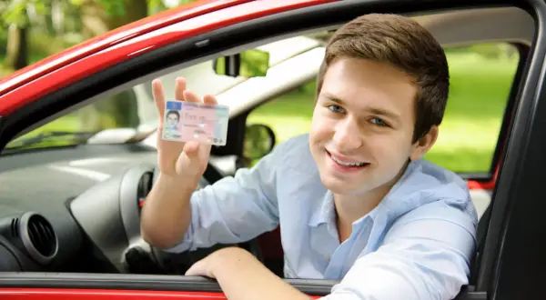 Uśmiechnięty młody mężczyzna w czerwonym samochodzie pokazuje prawo jazdy. Czy na prawo jazdy można wziąć kredyt na wymarzone auto?