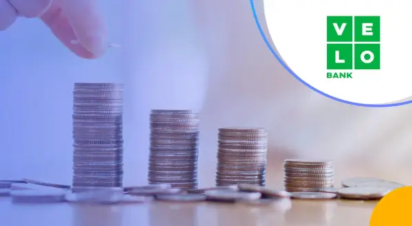Ręka dodaje monetę do rosnącej stosu, symbolizując wzrost kapitału. VeloBank oprocentowanie lokat rośnie wraz z Twoimi oszczędnościami.