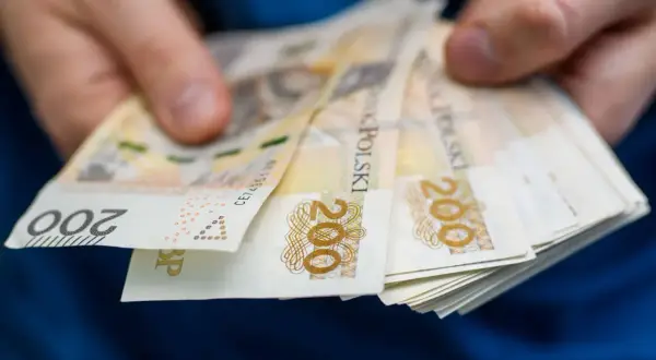 Dłonie trzymają plik banknotów 200 zł. Może to symbolizować wsparcie finansowe, np. stały zasiłek dla bezrobotnych po 55 roku życia.