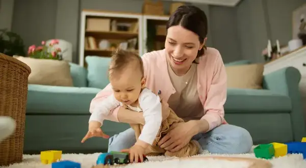 Uśmiechnięta mama bawi się z dzieckiem na dywanie, wśród kolorowych zabawek.