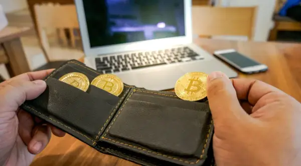 Ręka trzymająca portfel z trzema złotymi monetami bitcoin. Na stole leży laptop i smartfon. To symbol inwestycji na zonda giełda.