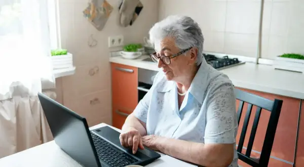 Starsza kobieta w okularach siedzi przy stole w kuchni i korzysta z laptopa.