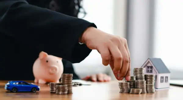 Dłoń układa monety obok skarbonki, modelu domu i samochodu.