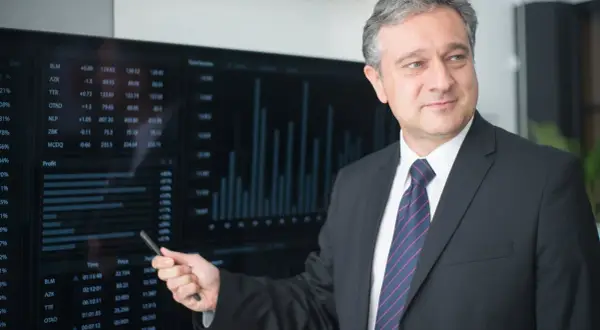 Mężczyzna w garniturze wskazuje na ekran z wykresami i danymi finansowymi.