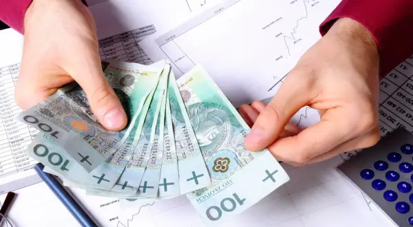 Dłonie trzymające stos polskich banknotów 100 zł na tle dokumentów finansowych i kalkulatora.