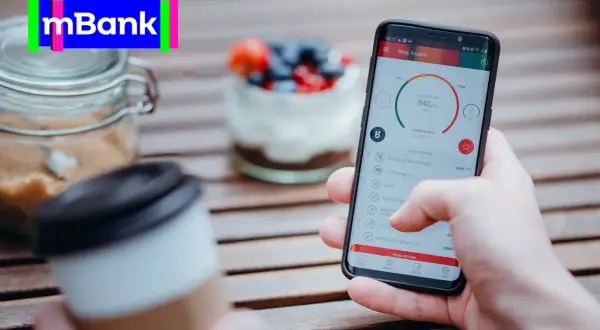 Dłoń trzyma smartfon z aplikacją mBank, na stole obok kawa i słoik z cukrem.
