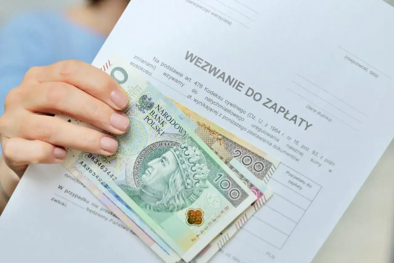 Dłoń trzyma wezwanie do zapłaty i polskie banknoty. Zastanawiasz się, po jakim czasie komornik wchodzi na konto? Dokumenty i pieniądze sugerują pilną potrzebę uregulowania długu.