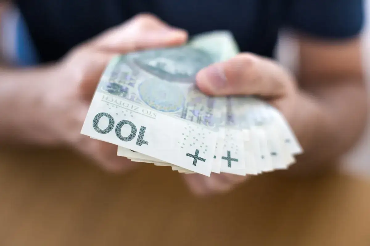Dłonie trzymają stos banknotów 100 zł. Może to symbolizować pomoc w spłacie zobowiązań, np. poprzez konsolidację zadłużenia.