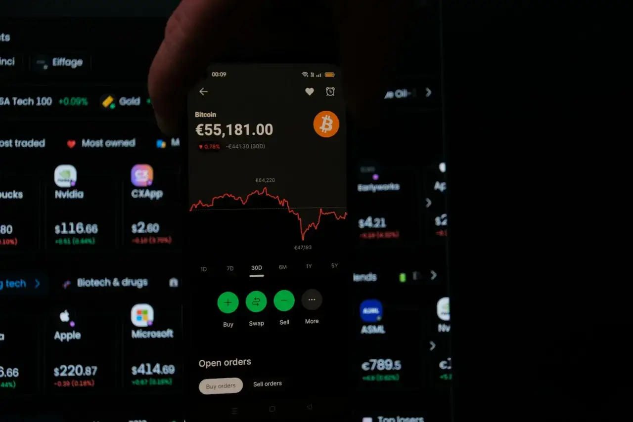 Na ekranie telefonu widać wykres Bitcoina, który pokazuje jego zmienność na giełdzie. Obok inne akcje i kryptowaluty.