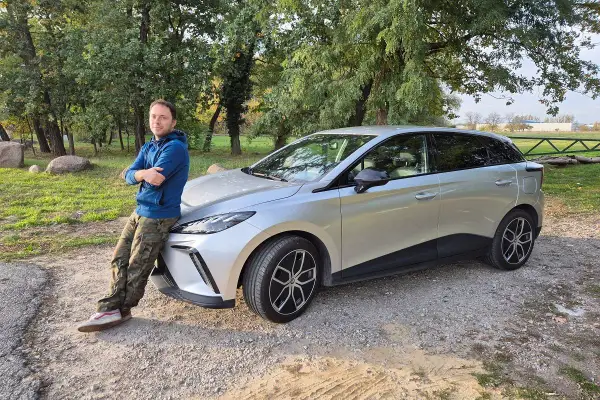 Mężczyzna opiera się o srebrny samochód, zastanawiając się, dlaczego nie warto kupować nowego samochodu.
