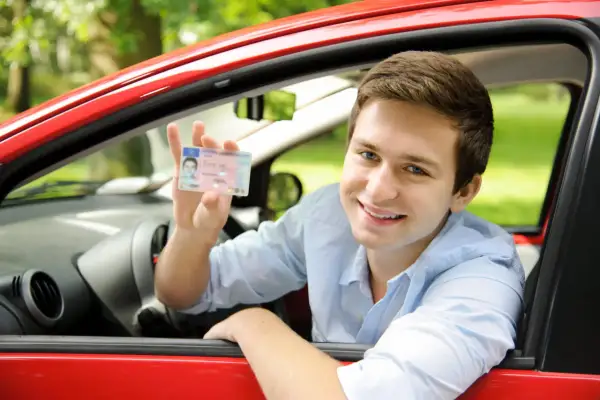 Uśmiechnięty młody mężczyzna w czerwonym samochodzie pokazuje prawo jazdy. Czy na prawo jazdy można wziąć kredyt na wymarzone auto?