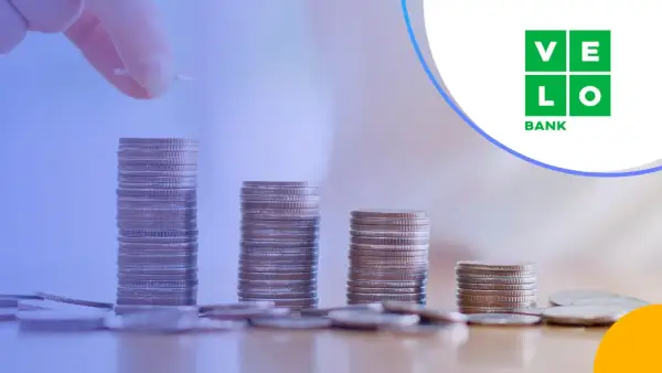 Ręka dodaje monetę do rosnącej stosu, symbolizując wzrost kapitału. VeloBank oprocentowanie lokat rośnie wraz z Twoimi oszczędnościami.
