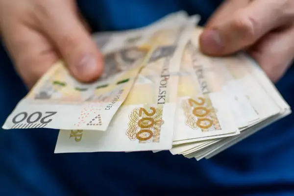 Dłonie trzymają plik banknotów 200 zł. Może to symbolizować wsparcie finansowe, np. stały zasiłek dla bezrobotnych po 55 roku życia.