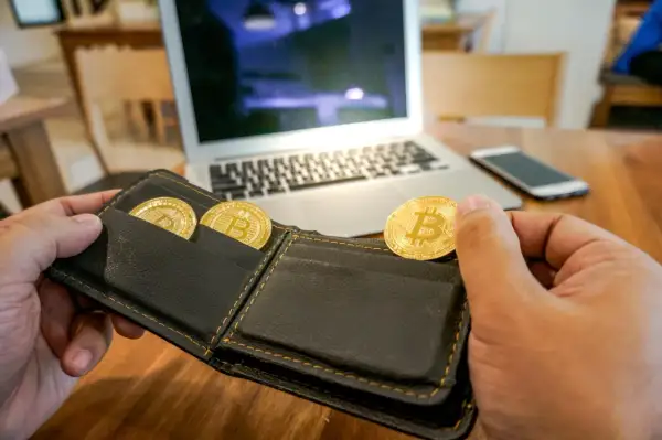 Ręka trzymająca portfel z trzema złotymi monetami bitcoin. Na stole leży laptop i smartfon. To symbol inwestycji na zonda giełda.