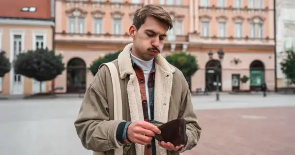 Młody mężczyzna z miną zmartwienia przegląda pusty portfel. W tle kamienice, symbolizujące inflację stylu życia.