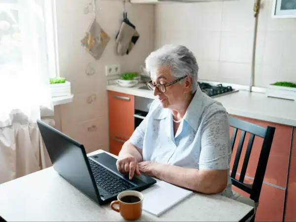 Starsza kobieta w okularach siedzi przy stole w kuchni i korzysta z laptopa.