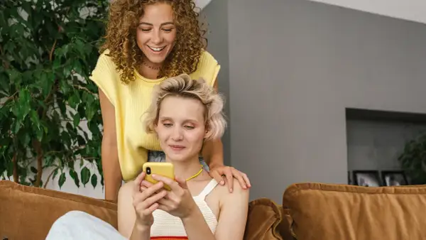 Dwie kobiety śmieją się, przeglądając telefon. Jedna z nich kładzie dłoń na ramieniu drugiej, sugerując wspólne konto dla par.