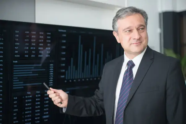 Mężczyzna w garniturze wskazuje na ekran z wykresami i danymi finansowymi.