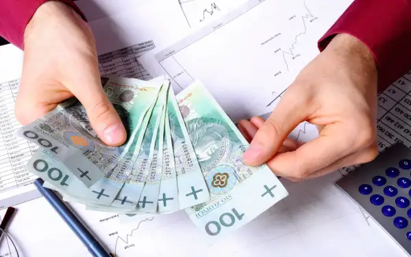 Dłonie trzymające stos polskich banknotów 100 zł na tle dokumentów finansowych i kalkulatora.
