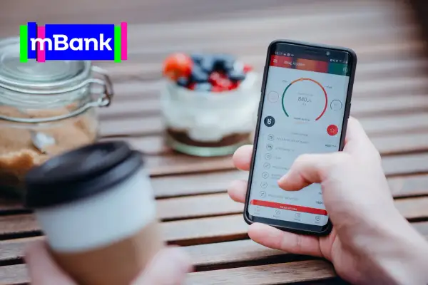 Dłoń trzyma smartfon z aplikacją mBank, na stole obok kawa i słoik z cukrem.